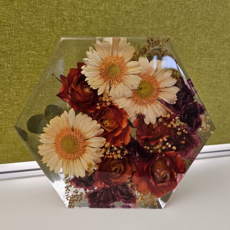 Eingeharzte Jubiläumsblumen in sechseckiger Form – helle Gänseblümchen, rote Rosen und Trockenblumen kunstvoll konserviert in klarem Harz.