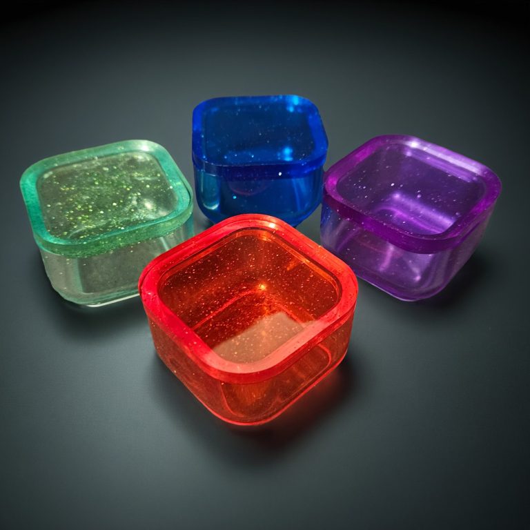 Vier kleine, viereckige Kästchen aus transparentem, glitzerndem Harz in Rot, Blau, Lila und Mintgrün.