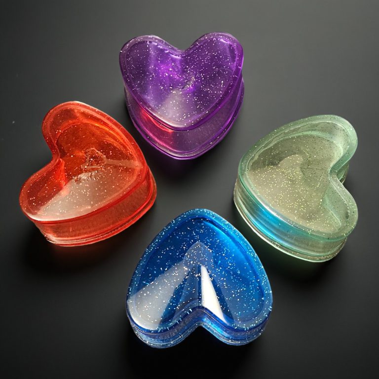 Vier herzförmige Harzkästchen mit Deckel in den Farben Rot, Blau, Grün und Lila – transparent mit Glitzereffekt, aus Epoxidharz.